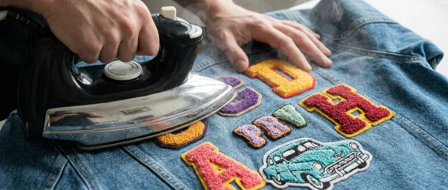 Person using a clothes iron to apply colorful chenille letter patches and a car patch onto a denim jacket