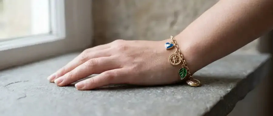 A close-up wrist shot of a hand resting casually on a neutral stone or marble surface, wearing a curated charm bracelet