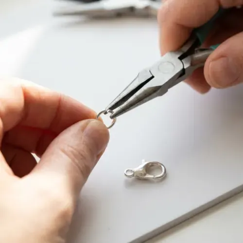 Close-up of hands using chain-nose pliers to twist open a silver jump ring sideways next to a silver lobster clasp on a white craft mat