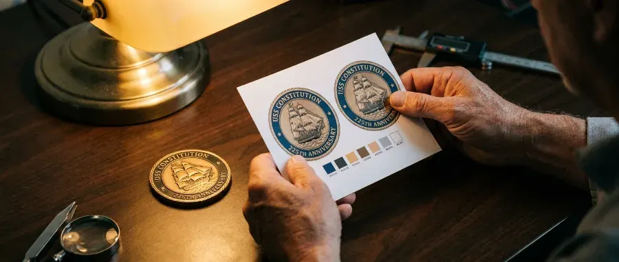 Person holding a printed challenge coin digital proof under desk lamp light with a finished physical coin beside it during the final review before approval