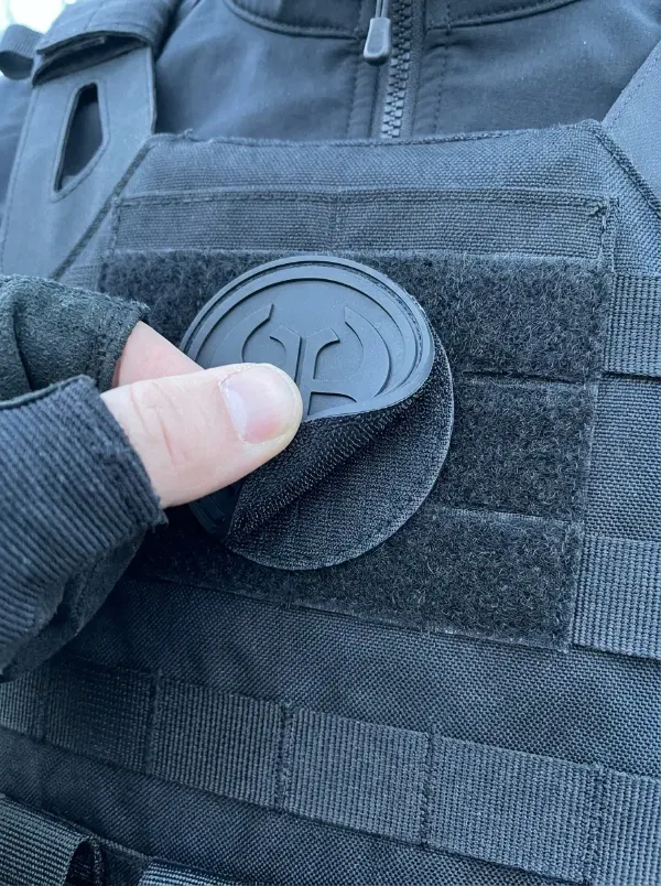 Hand pressing a custom PVC patch onto a tactical morale patch panel on a black plate carrier vest showing hook-and-loop backing connection