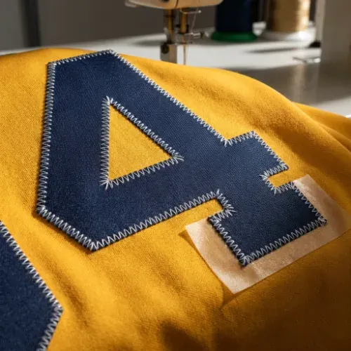 Close up of bold tackle twill lettering being stitched onto a team jersey with heat-resistant tape visible underneath